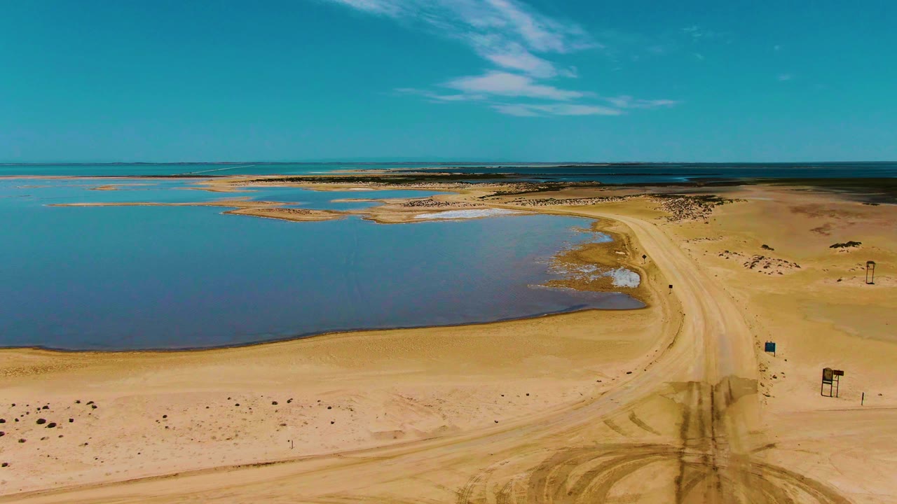 High aerial frame over Guerrero Negro, Baja California Sur, showing blue water on the left, a pale sandy road curving through the center, and no visible people.
