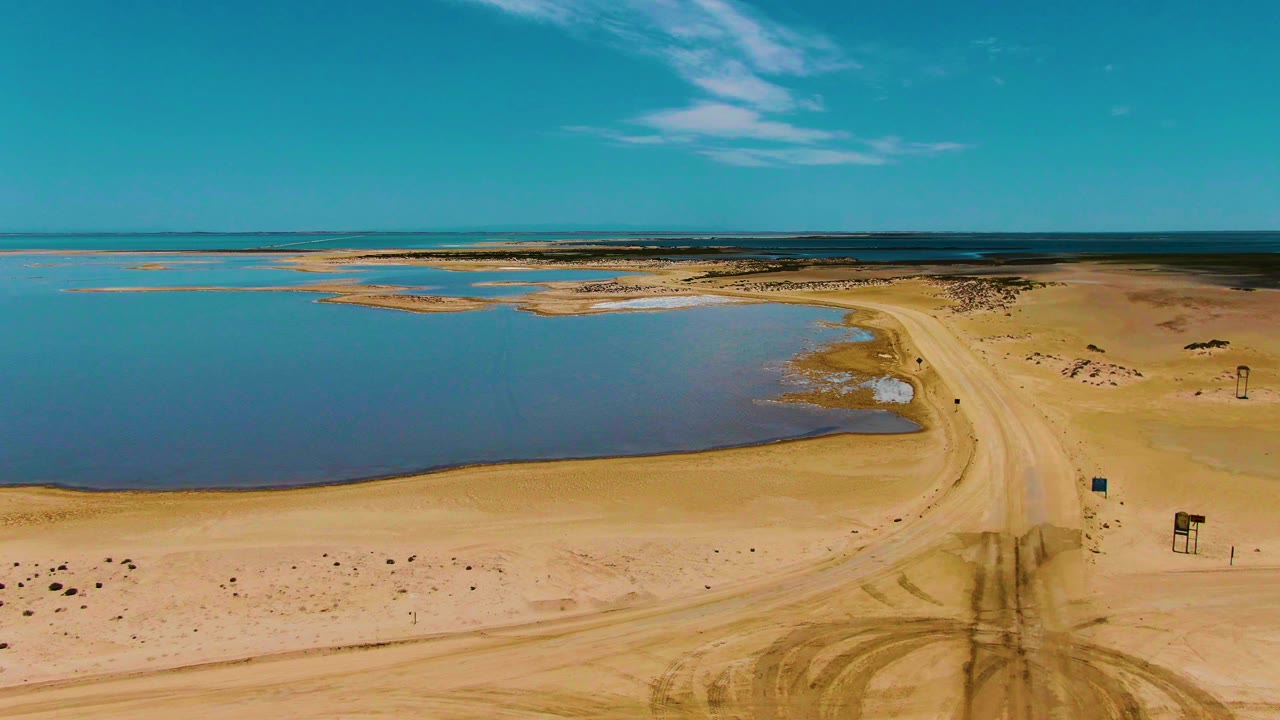 Wide aerial frame in Guerrero Negro, Baja California Sur, with calm blue water, a sandy road, dark tire tracks in the foreground, and no visible people.
