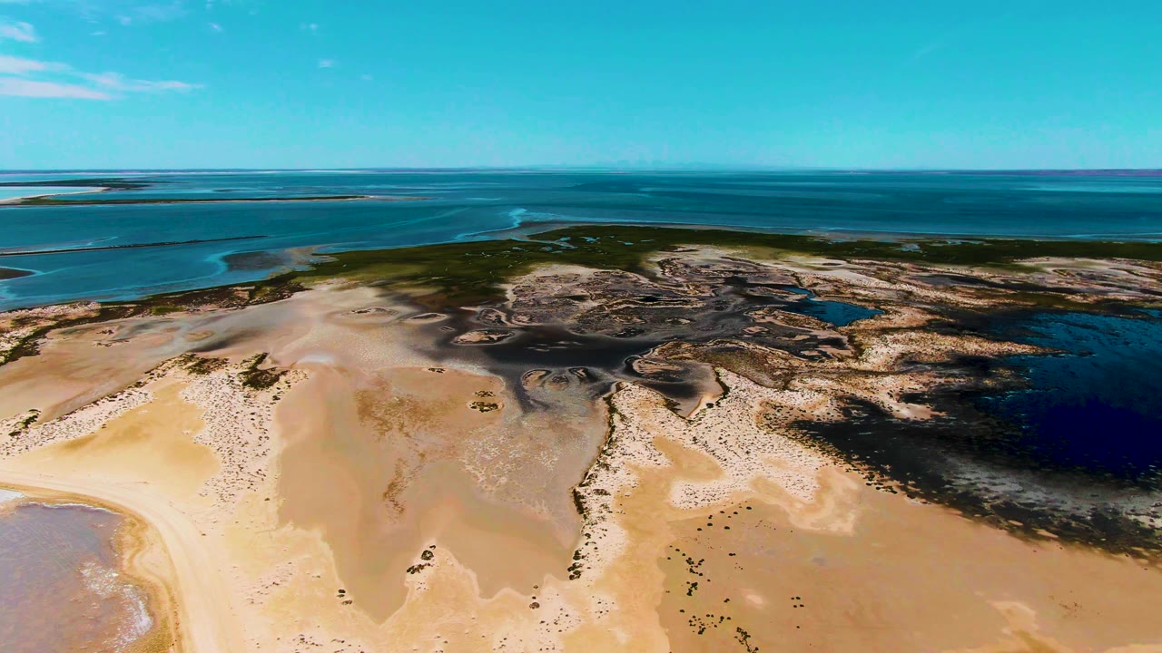 Aerial frame over Guerrero Negro tidal flats with pale sand in front, dark wet channels near the center, blue ocean on the horizon, and no visible people.