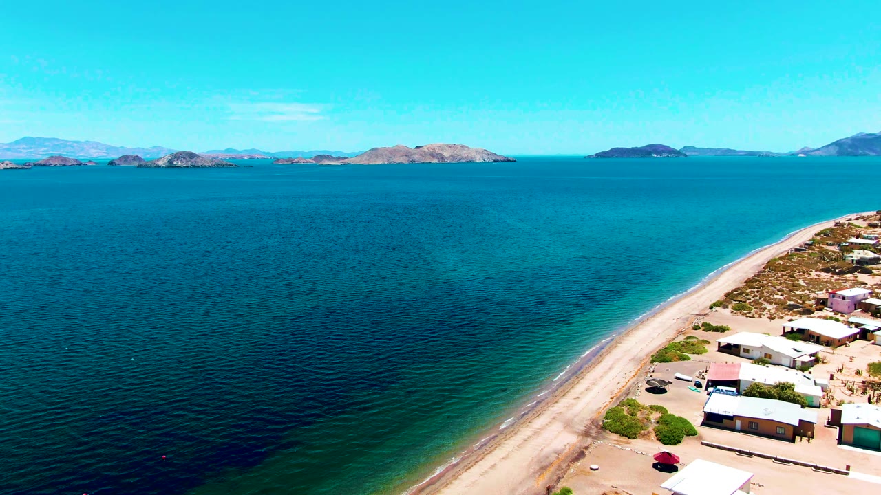 Aerial frame over Bahia De Los Angeles showing a sandy shore curving past small coastal buildings, calm blue water, distant islands, mountains, and no visible people.
