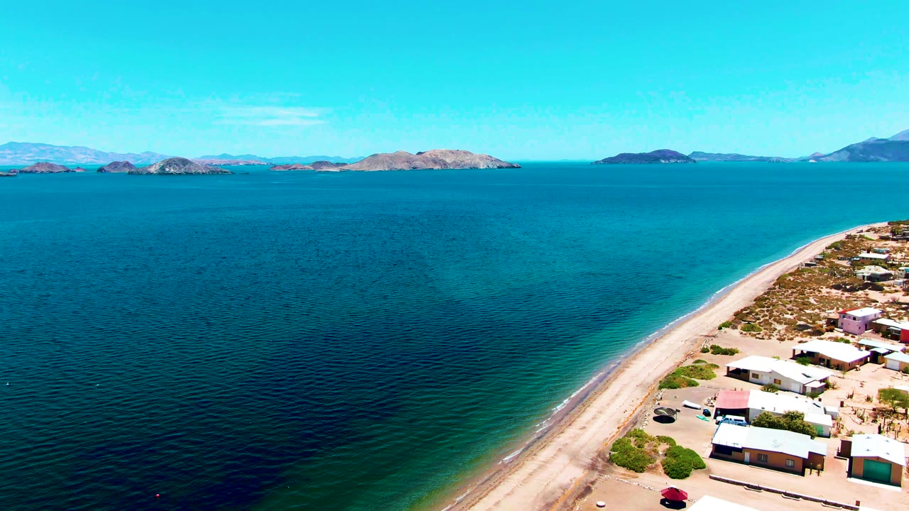 High aerial view of turquoise water beside a sandy beach road and small shore buildings.