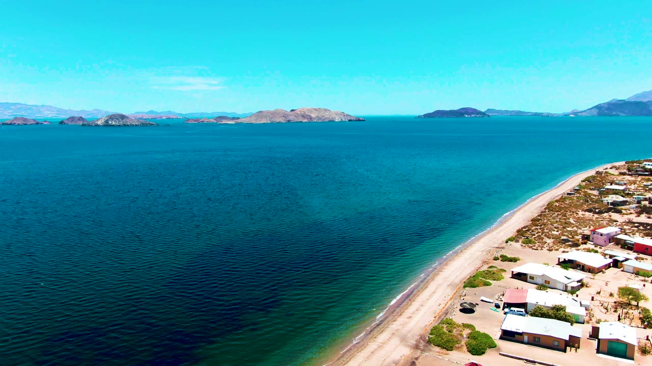 Aerial frame over Bahia De Los Angeles showing calm bay water across most of the image, a curved sandy shoreline with small buildings to the right, distant islands, and no visible people.