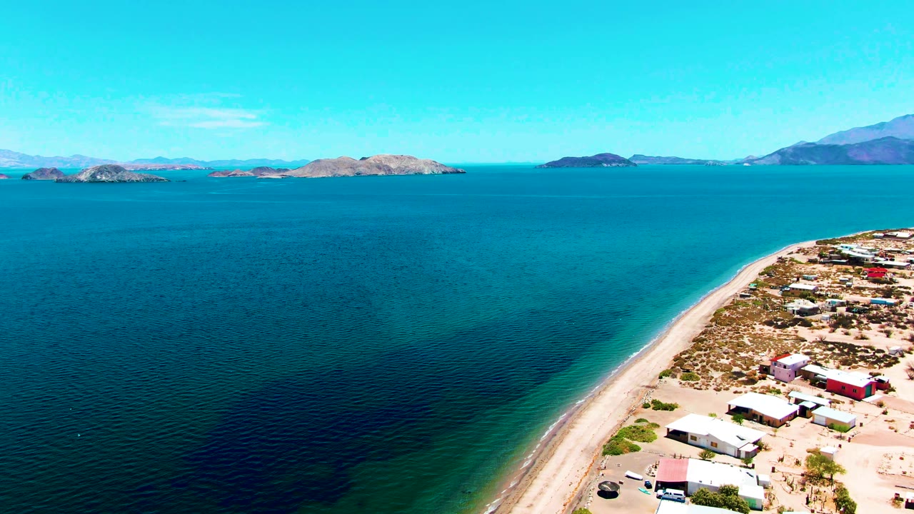 Aerial frame over Bahia De Los Angeles shows deep blue water, a diagonal sandy beach, small coastal buildings on the right, distant islands, mountains, and no visible people.