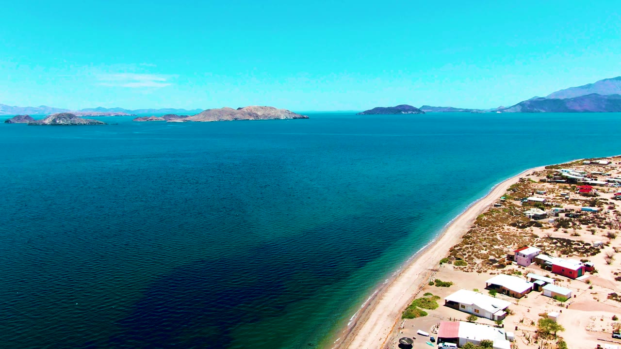 Drone view of calm water, sandy shore, scattered buildings, islands, and mountains.