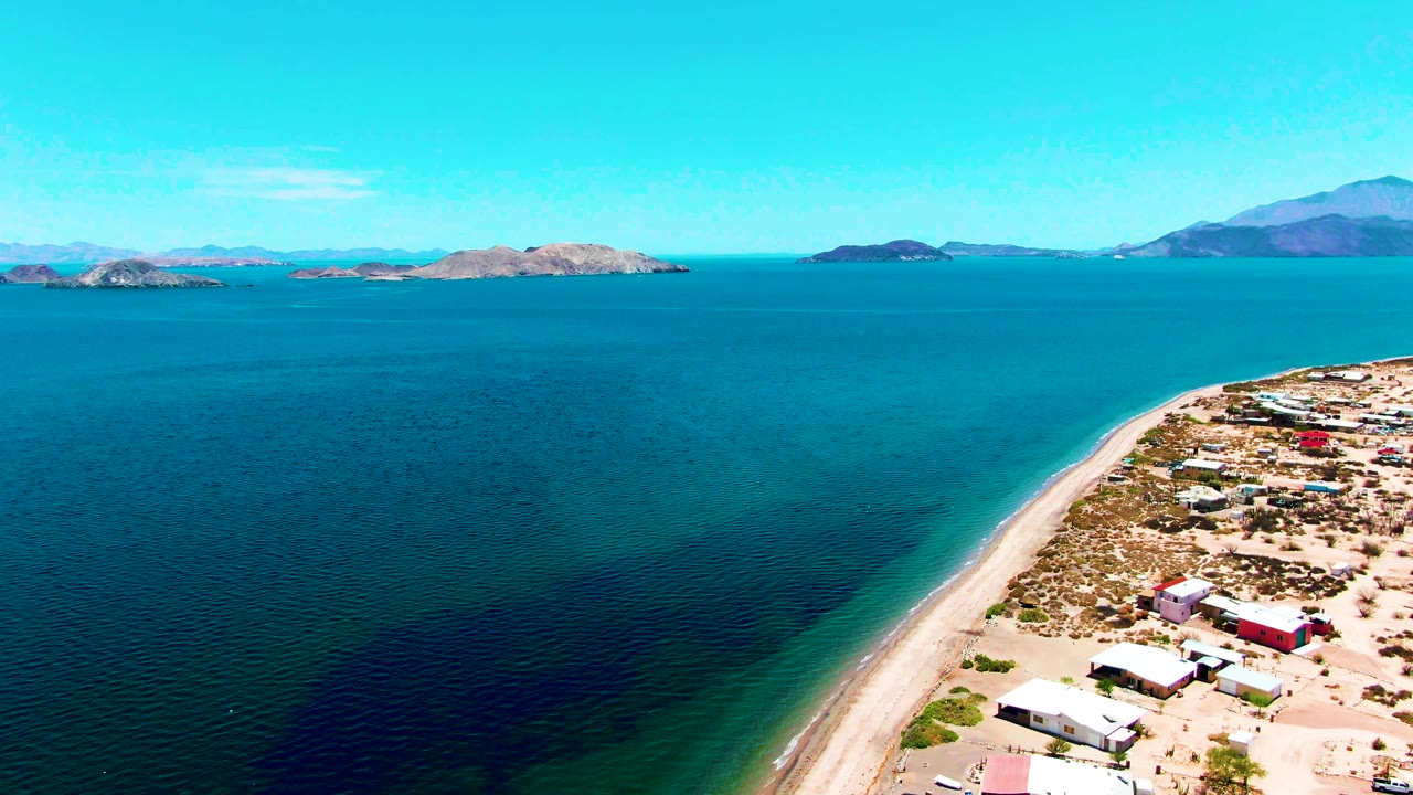 At 0:02, the drone frame shows turquoise shallows along the beach, darker patches in the water, low coastal buildings to the right, distant rocky islands, mountains, and no visible people.