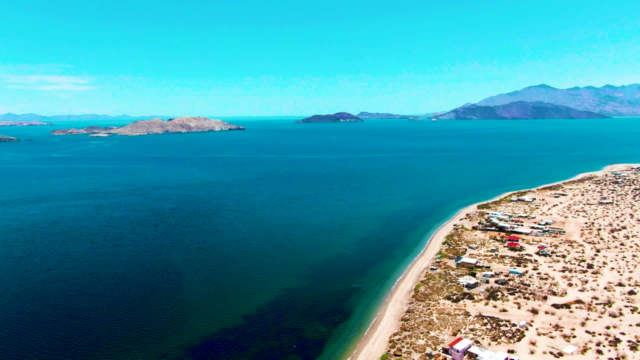 Aerial frame over Bahia De Los Angeles shows a curving sandy beach with scattered shoreline structures, a small red vehicle, blue ocean, distant islands, mountains, and no visible people.