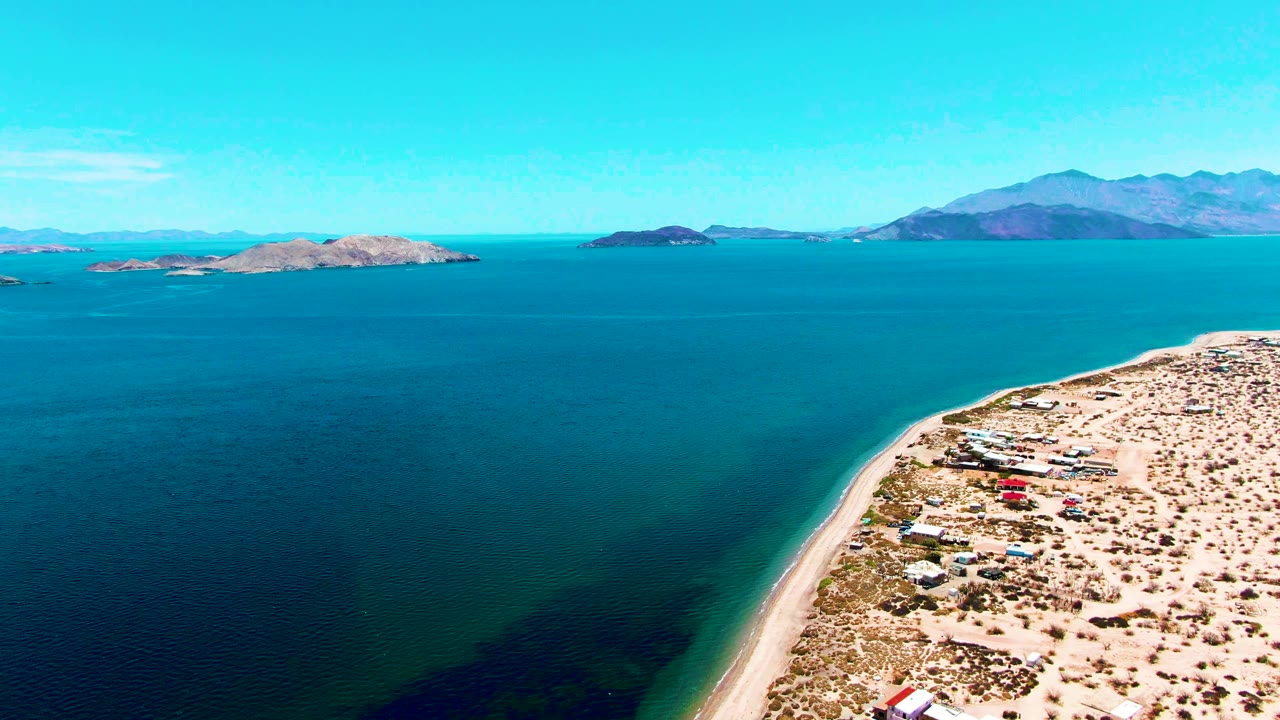 Wide aerial coastline with turquoise water, sandy beach edge, islands, and mountains.