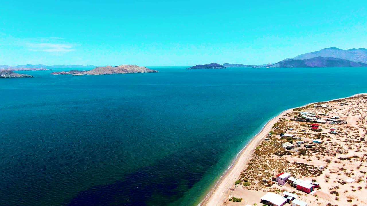 A wide aerial frame over Bahia De Los Angeles shows blue ocean, a sandy shoreline with small beach structures, distant islands, mountains on the horizon, and no visible people.