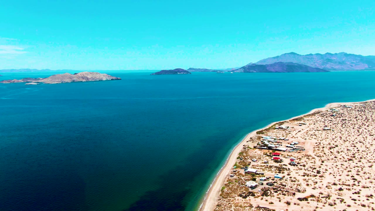 Aerial frame over Bahia De Los Angeles showing pale sandy shoreline, sparse desert scrub, small coastal structures, blue bay water, distant islands, mountains, and no visible people.
