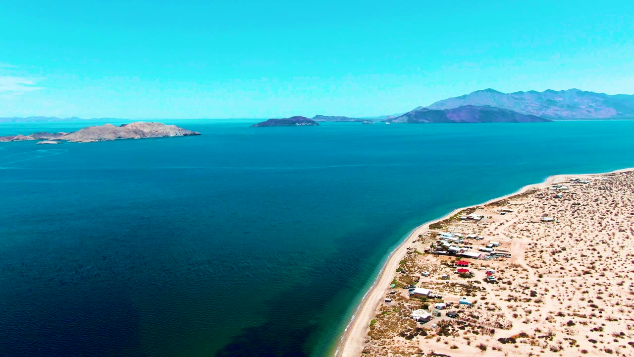 Wide aerial coastline with turquoise water, beach edge, desert terrain, islands, and mountains.