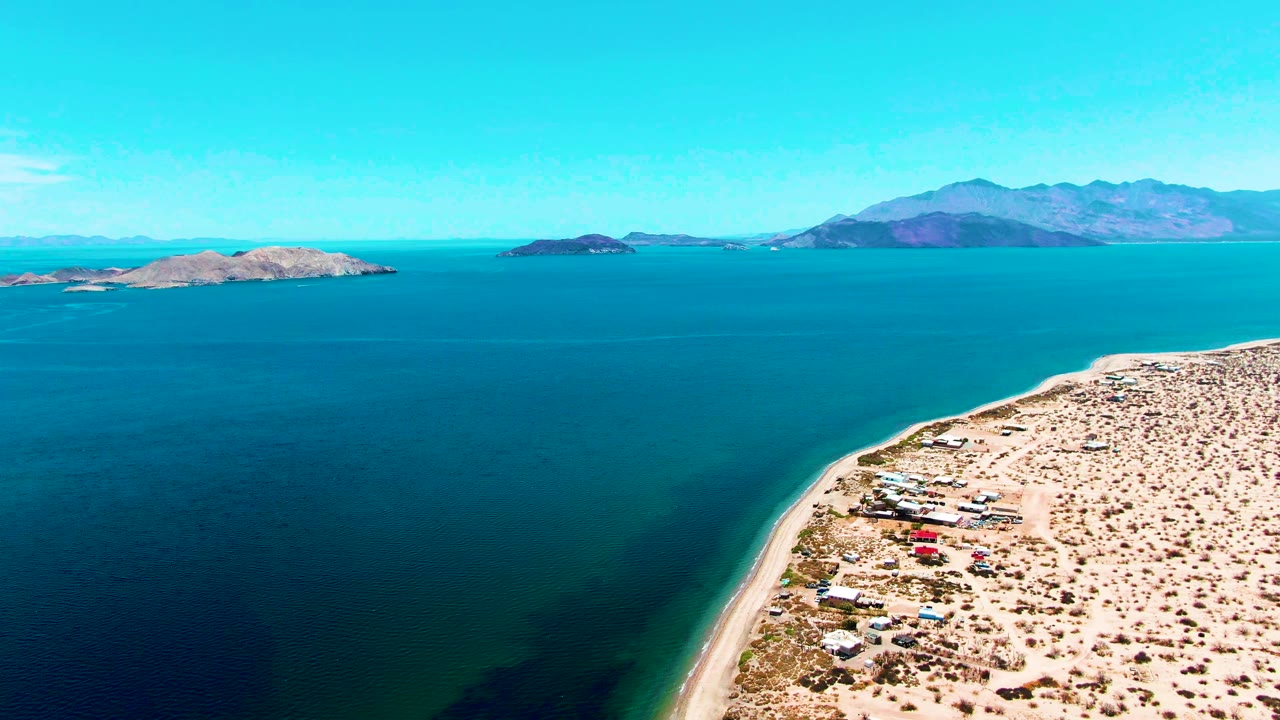 At 0:02, the aerial view shows more open water across the center of Bahia De Los Angeles, with sandy coast at lower right, clustered small structures, low islands, distant mountains, and no visible people.
