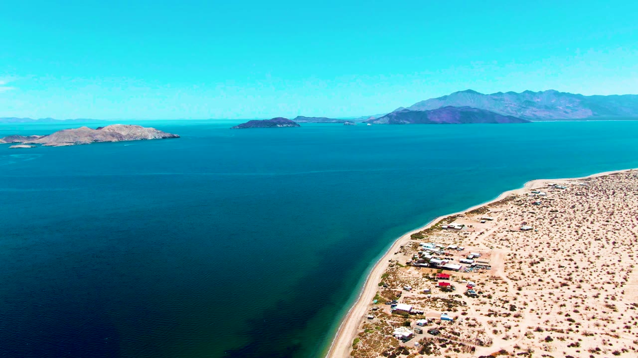 Aerial frame over Bahia De Los Angeles showing turquoise water, a sandy shore path, arid land at right, distant islands and mountains, with no visible people.