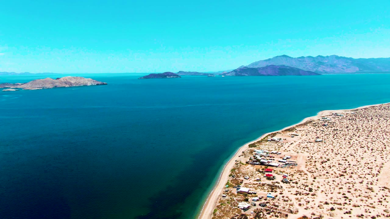 Aerial view in Bahia De Los Angeles with calm blue water filling the frame, sandy coast and scattered structures at lower right, and no visible people.