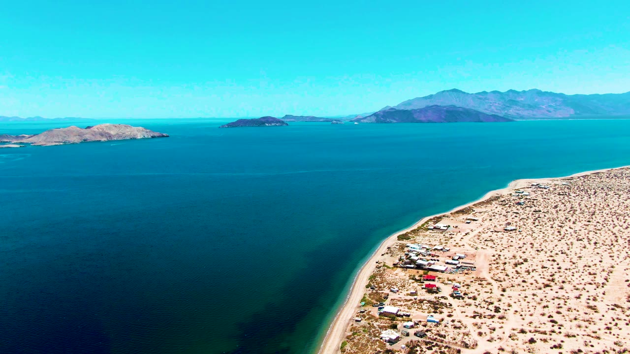 Bahia De Los Angeles aerial frame with a curved sandy shoreline, dark shallow water near the beach, open blue sea, islands, mountains, and no visible people.