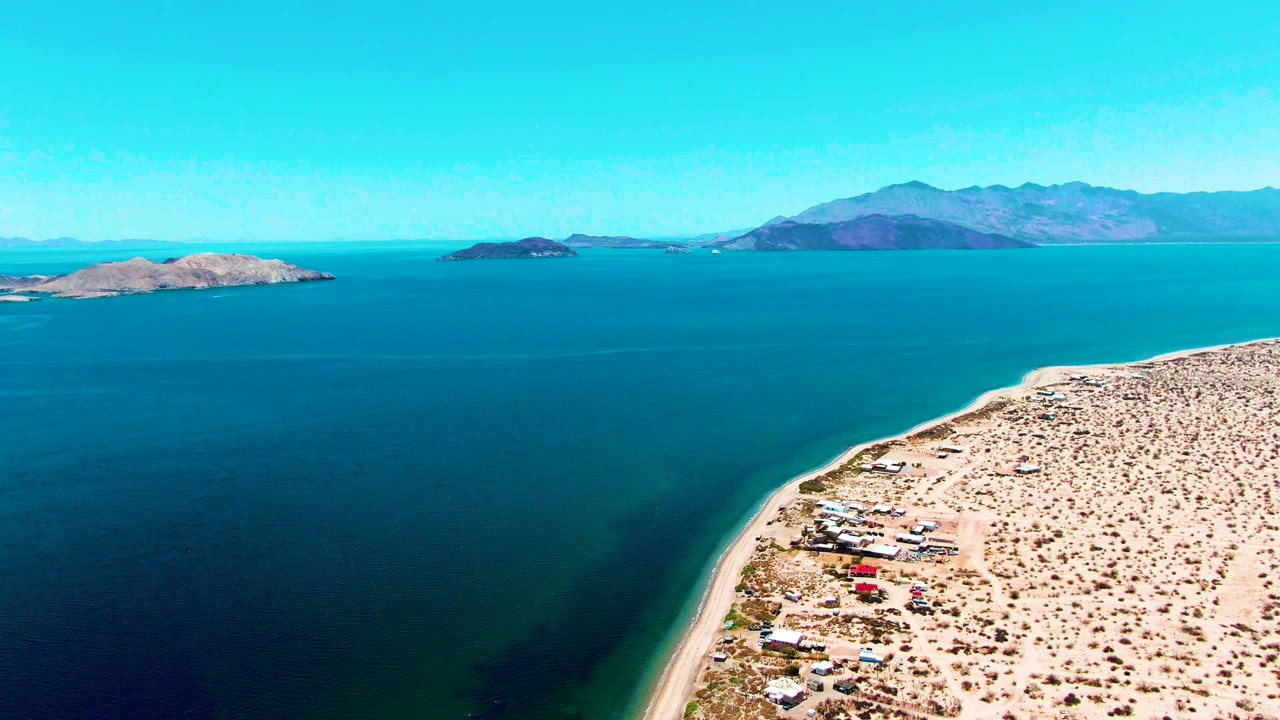 Wide aerial over blue ocean with desert coast at right and islands on the horizon.