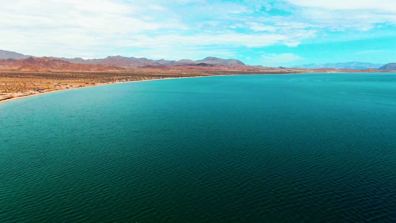 Opening frame over Bahia De Los Angeles shows turquoise water in the foreground, a sandy left shoreline, desert flats, distant mountains, scattered clouds, and no visible people.