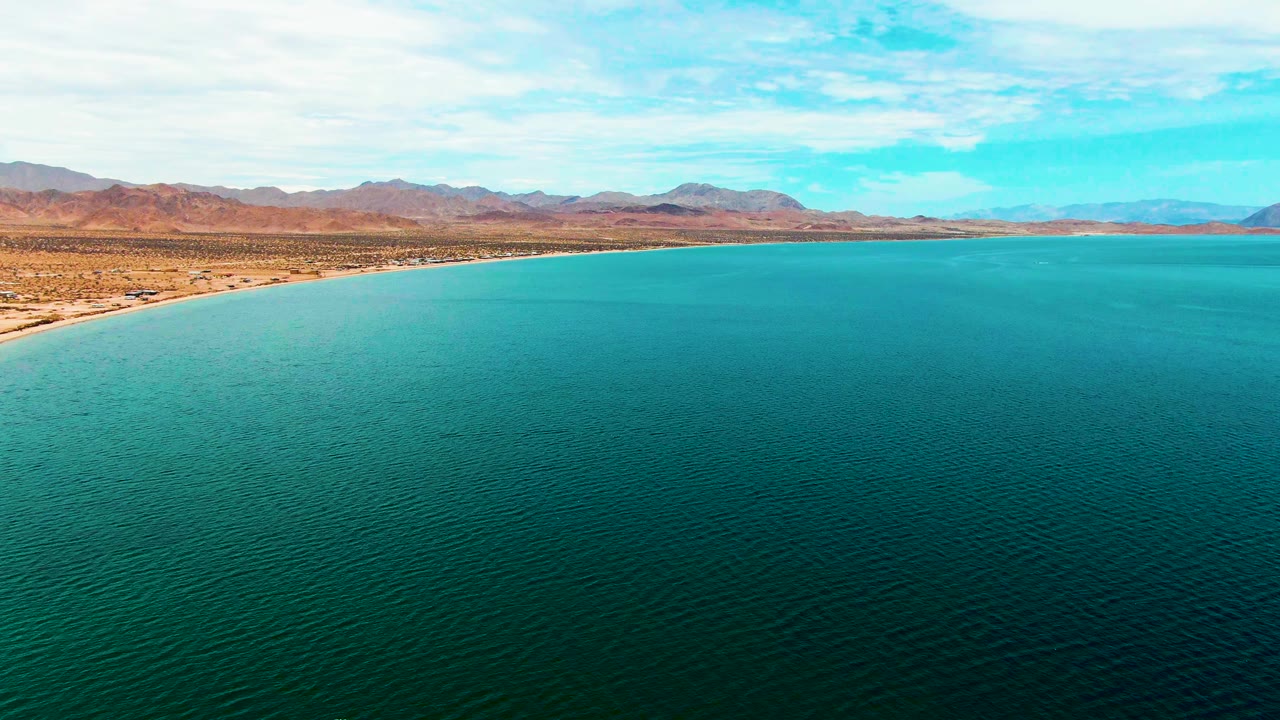 At 0:01, Bahia De Los Angeles appears as a broad turquoise bay with the beach curving from the left edge, dry terrain behind it, distant mountains, high clouds, and no visible people.