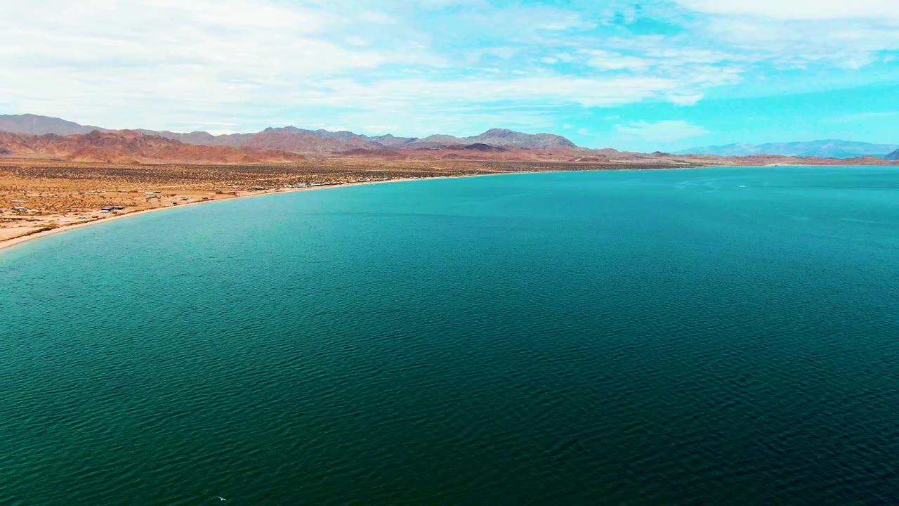 At 0:02, the frame holds on Bahia De Los Angeles with darker turquoise water across the foreground, a thin sandy shore at left, desert flats, a low mountain range, and no visible people.