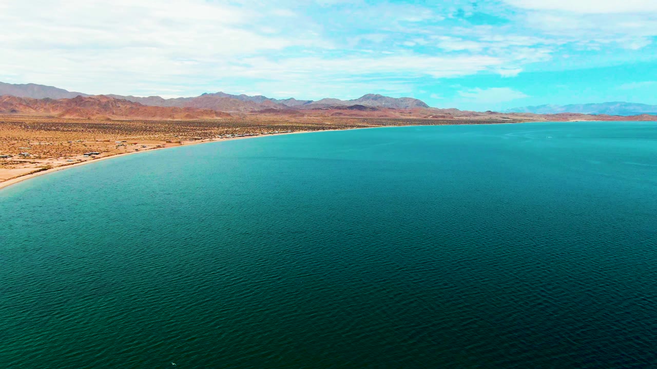 Calm turquoise water meets a sandy beach with desert land and mountains under clouds.