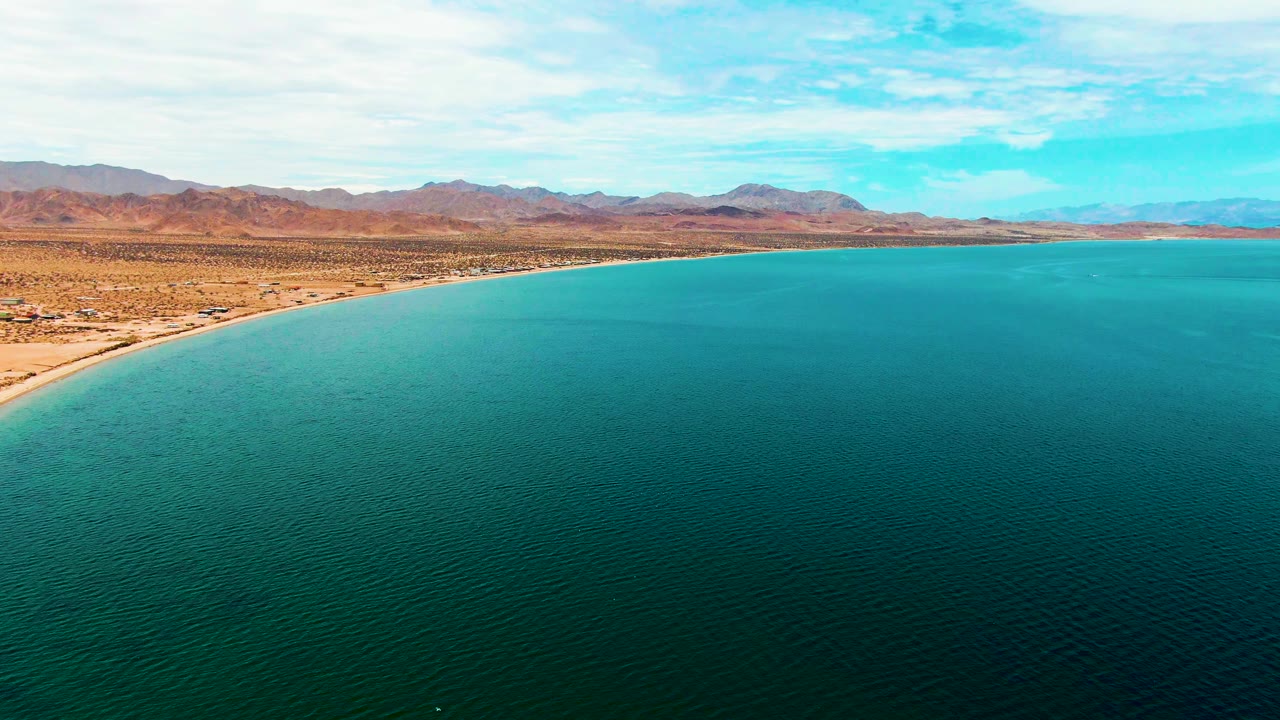 Wide aerial view over Bahia De Los Angeles with turquoise ocean in the foreground, a sandy beach curving along arid desert, distant mountains, blue sky, and no visible people.