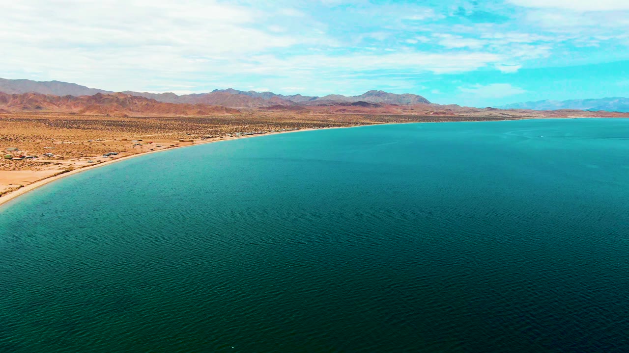 Aerial frame of Bahia De Los Angeles showing a long sandy shoreline between deep turquoise water and dry desert terrain, with low mountains under thin clouds and no visible people.