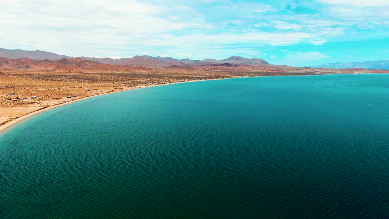 High aerial view in Bahia De Los Angeles with broad turquoise water, a pale beach edge, scrubby desert flats, reddish-brown mountains, scattered clouds, and no visible people.