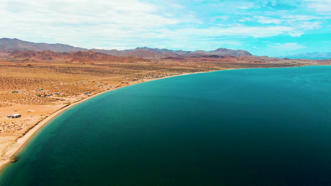 Aerial view over Bahia De Los Angeles with turquoise water on the right, sandy desert shore on the left, distant brown mountains, partly cloudy sky, and no visible people.
