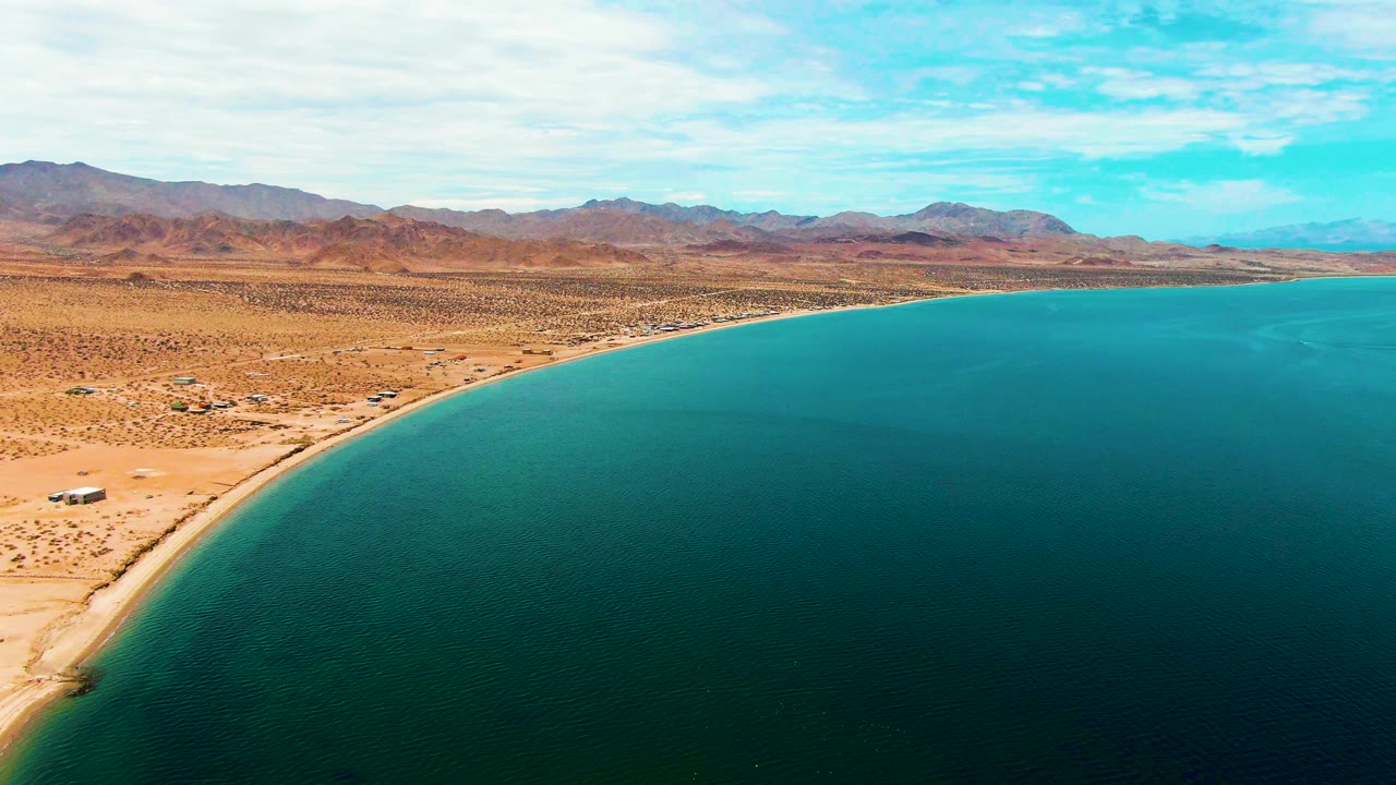 Bahia De Los Angeles coastline curves through the frame, with deep turquoise water in the foreground, pale sand along the edge, desert scrub inland, distant mountains, and no visible people.