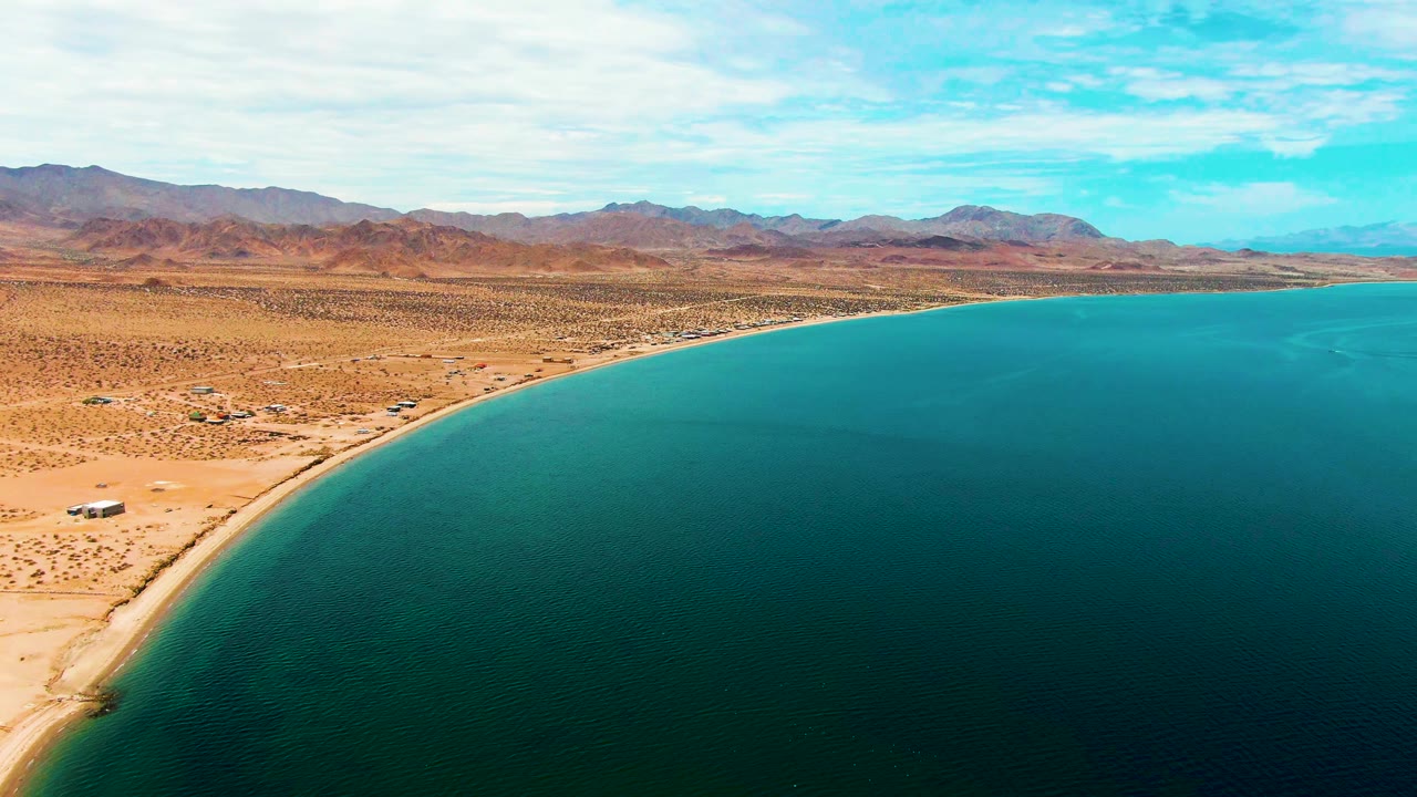 Wide aerial frame of Bahia De Los Angeles with open turquoise bay occupying the lower right, arid shoreline and desert flats to the left, brown mountains across the horizon, and no visible people.