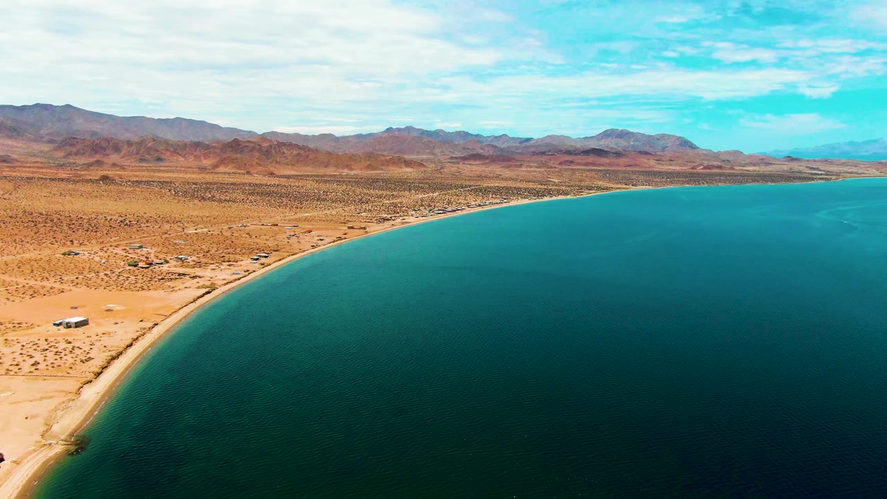 Aerial view along Bahia De Los Angeles where a narrow sandy beach borders turquoise water, with desert plain and low mountains behind it and no visible people.