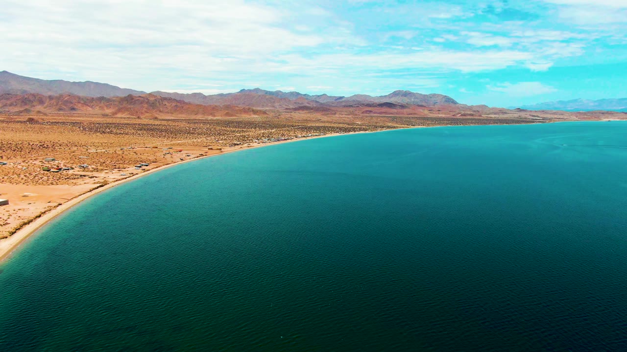 Aerial frame over Bahia De Los Angeles with turquoise water filling the foreground, a sandy curved shoreline at left, desert flats, distant mountains, scattered clouds, and no visible people.