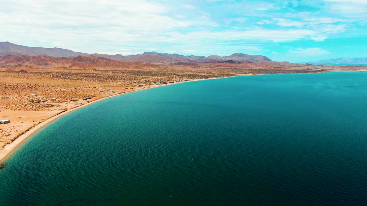 Wide aerial view in Bahia De Los Angeles showing a smooth arc of sand between turquoise water and tan desert plain, with mountains across the horizon and no visible people.