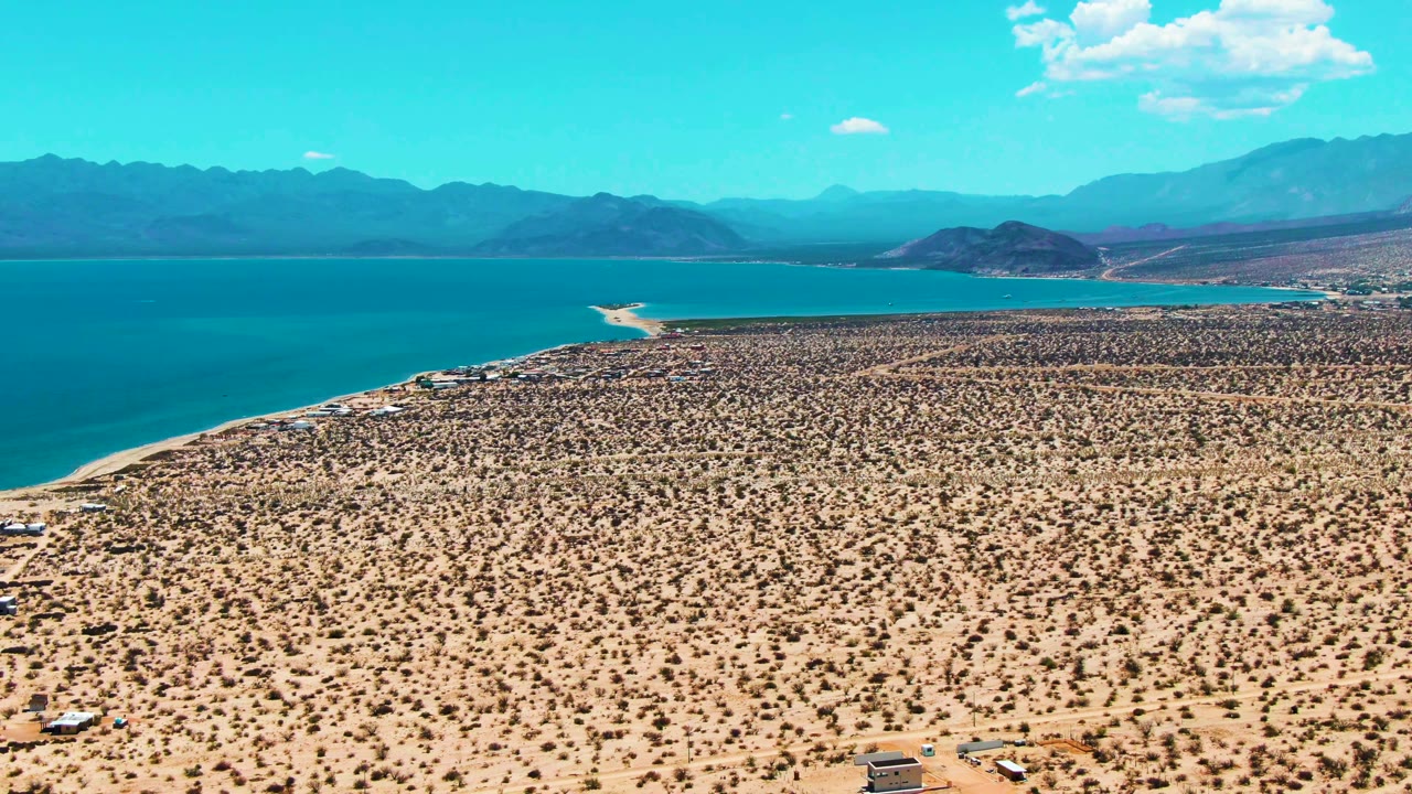 Opening aerial frame over Bahia De Los Angeles, with sandy desert scrub in the foreground, blue water beyond the shoreline, distant mountains on the horizon, and no visible people.