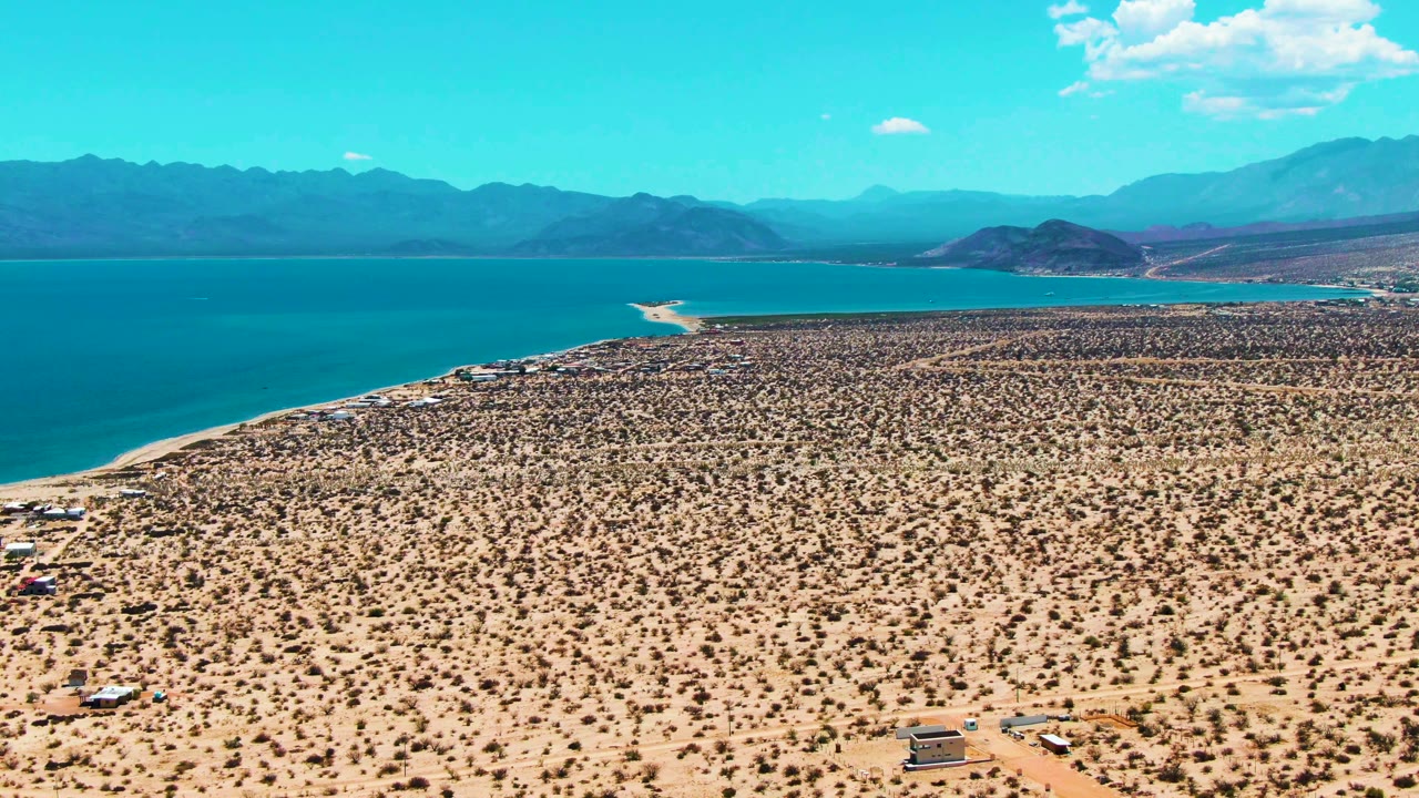High aerial landscape of sandy desert, blue water, low hills, and mountain range.