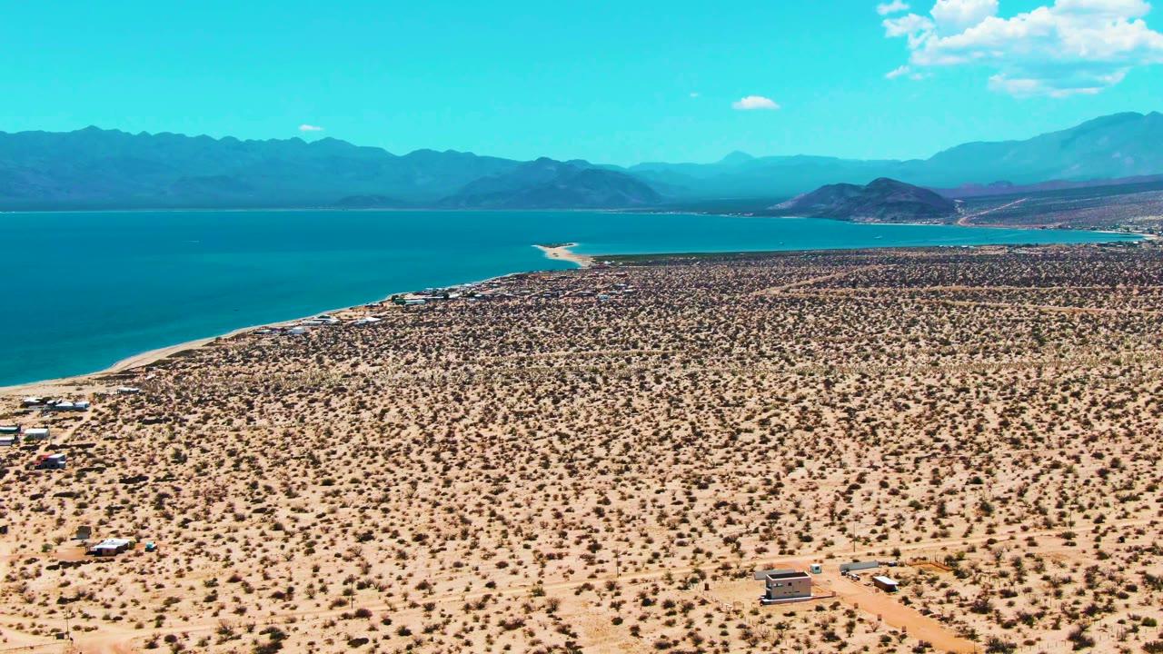 At 0:00, an aerial view over Bahia De Los Angeles shows sandy desert scrub in front, blue water to the left, distant mountains on the horizon, and no visible people.