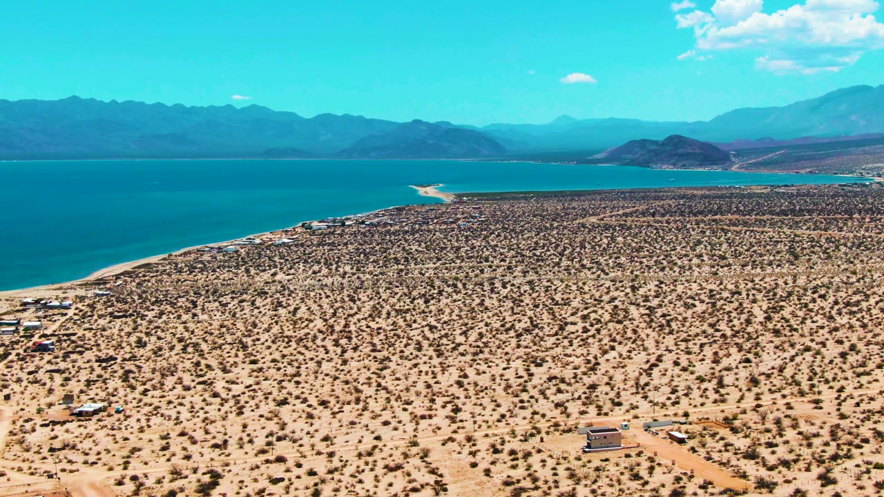 At 0:01, the aerial frame holds on Bahia De Los Angeles with a dirt road and scattered structures below, blue water beyond the desert plain, and no visible people.