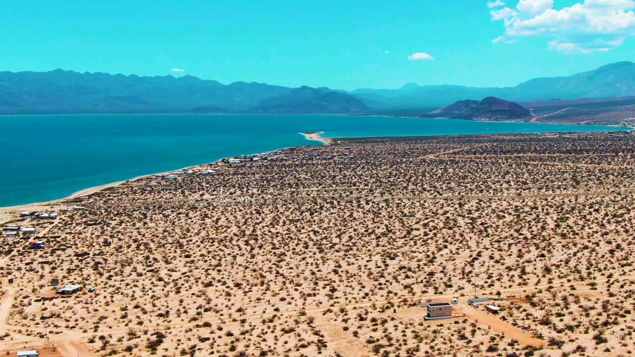 At 0:02, Bahia De Los Angeles appears from above with scrub-covered sand, faint dirt tracks, a curving shoreline, bright blue water, mountains, and no visible people.