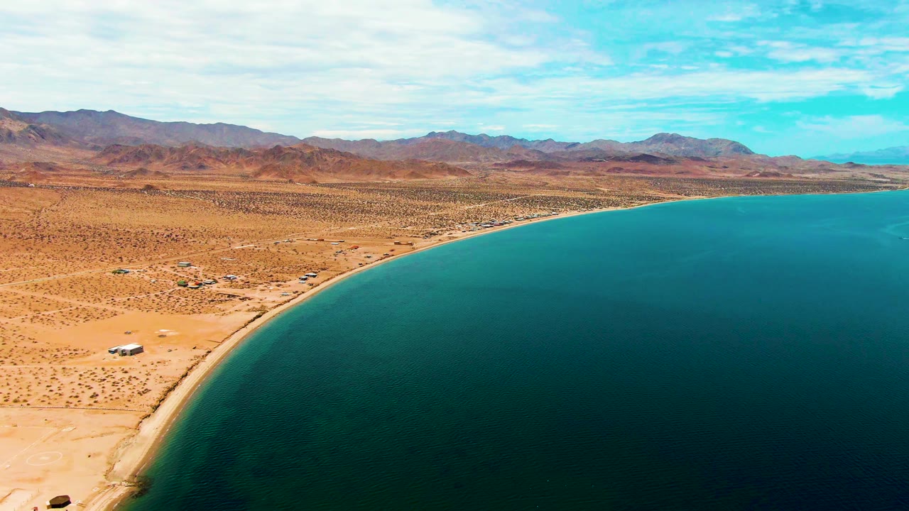 Aerial frame over Bahia De Los Angeles showing a curved sandy desert beach, deep blue water to the right, distant brown mountains, and no visible people.