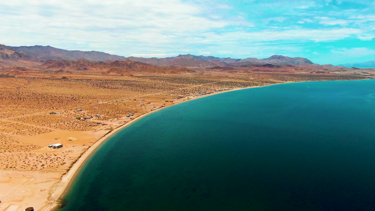 Aerial frame over Bahia De Los Angeles with a small white vehicle near the sandy shoreline, blue water filling the right side, desert terrain inland, and no visible people.