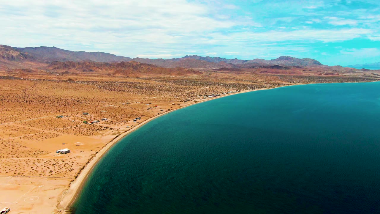 Aerial view of sandy beach, desert flats, blue water, and a white vehicle near the lower edge.