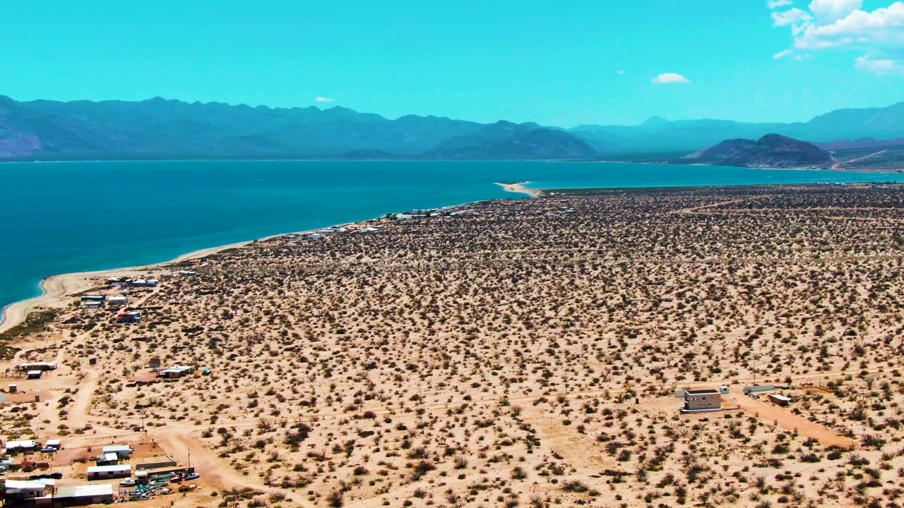 At 0:00, an aerial view over Bahia De Los Angeles shows sandy scrubland, a small desert camp, dirt roads, blue water, distant mountains, and no visible people.