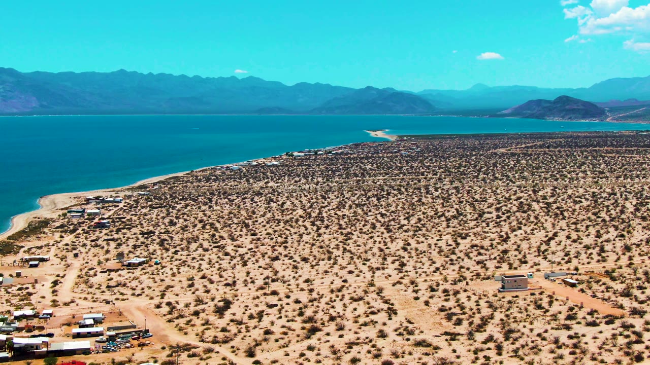At 0:01, the Bahia De Los Angeles aerial frame holds on scattered desert shrubs, low camp structures, pale dirt tracks, turquoise water, distant mountains, and no visible people.