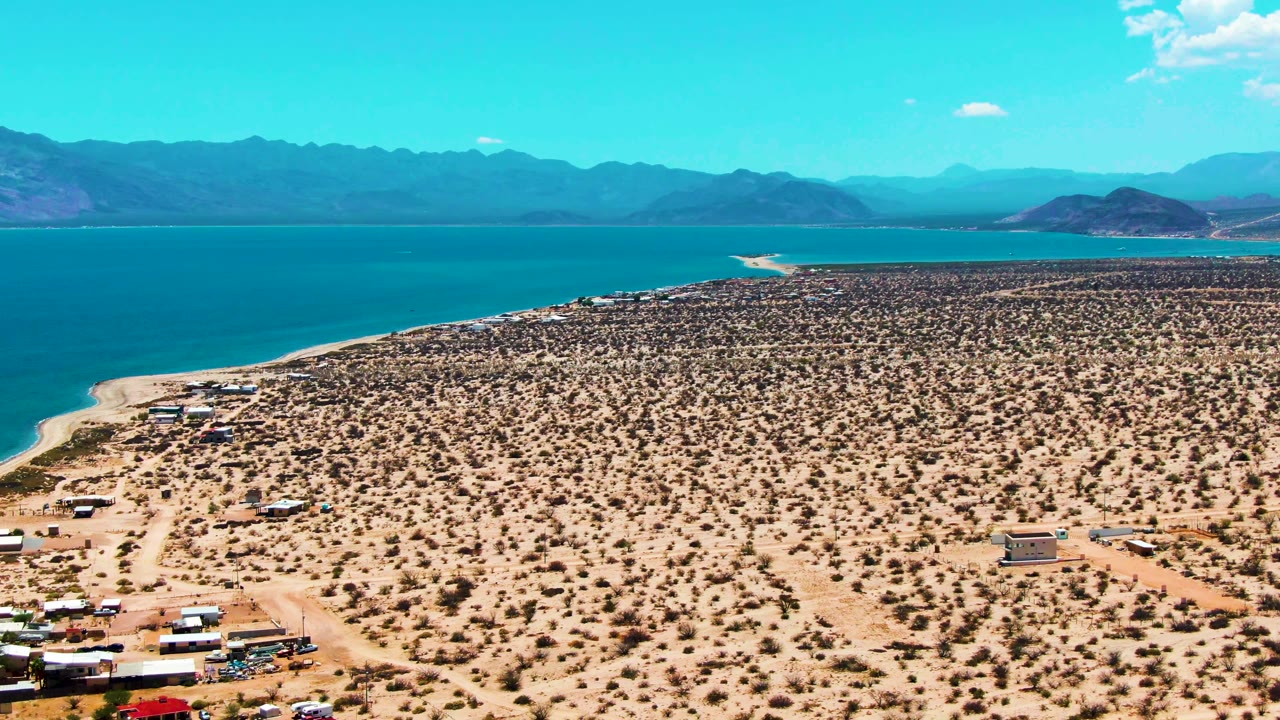 At 0:02, the high aerial composition shows a dirt road curving through a sparse camp area toward the blue water and mountains of Bahia De Los Angeles, with no visible people.