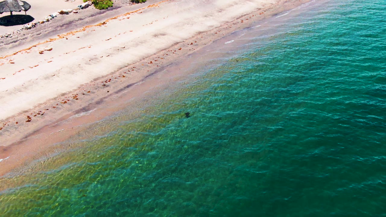 Mid-clip aerial view over Bahia De Los Angeles, with umbrellas at the top beach edge, rippled turquoise water below, and the distant wader still small in the shallows.