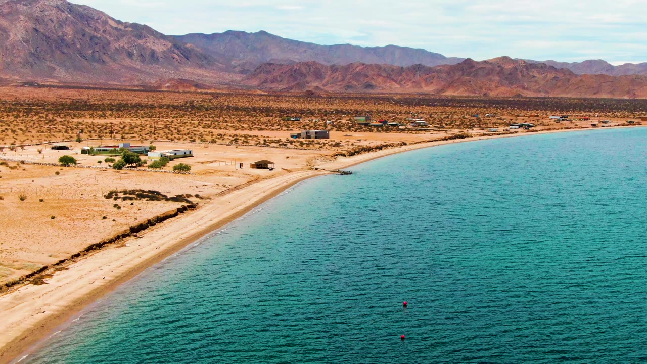 Aerial frame of Bahia De Los Angeles showing turquoise rippled water, a pale curving beach, sparse desert scrub, distant buildings, reddish mountains, and no visible people at the start of the clip.