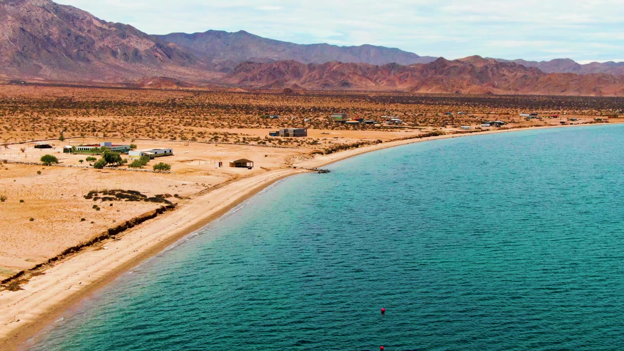 Second frame over Bahia De Los Angeles with the shoreline running diagonally across the image, offshore buoys lower in the water, scattered buildings on the sand, and no visible people.