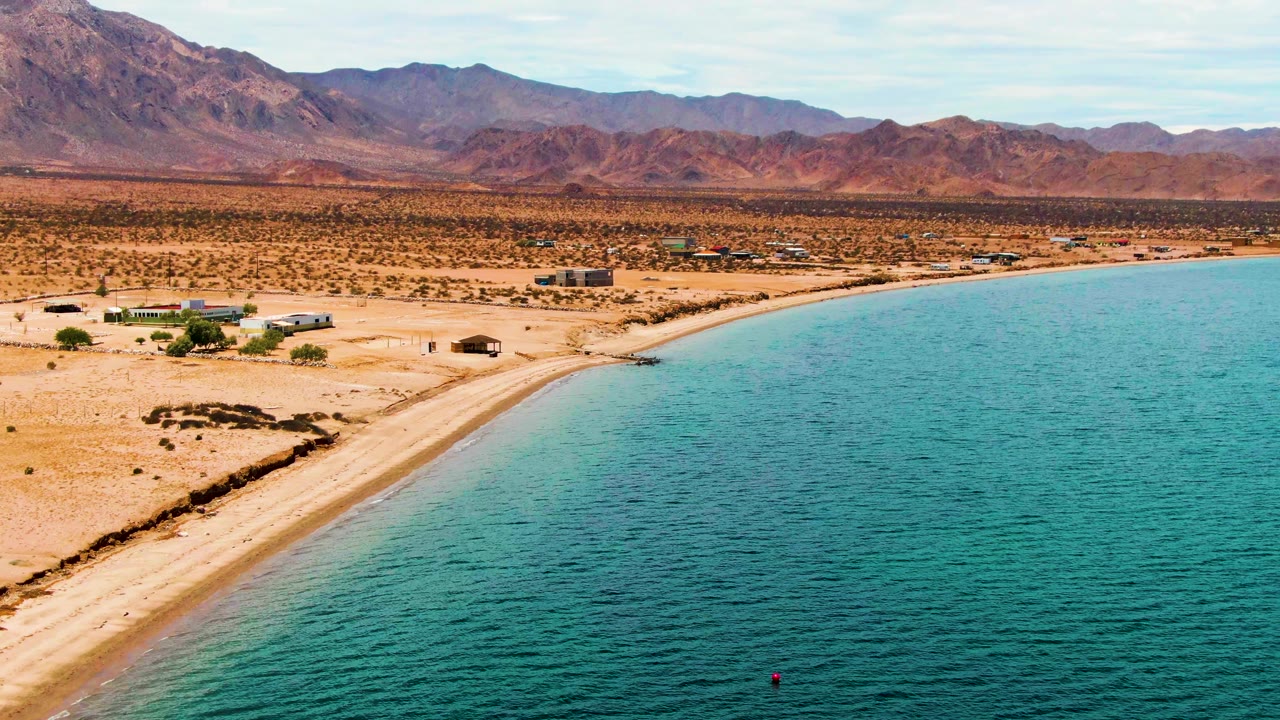 Aerial view of Bahia De Los Angeles with blue-green water filling the right side, a narrow sandy beach on the left, small shore structures, one red buoy near the lower frame, and no visible people.