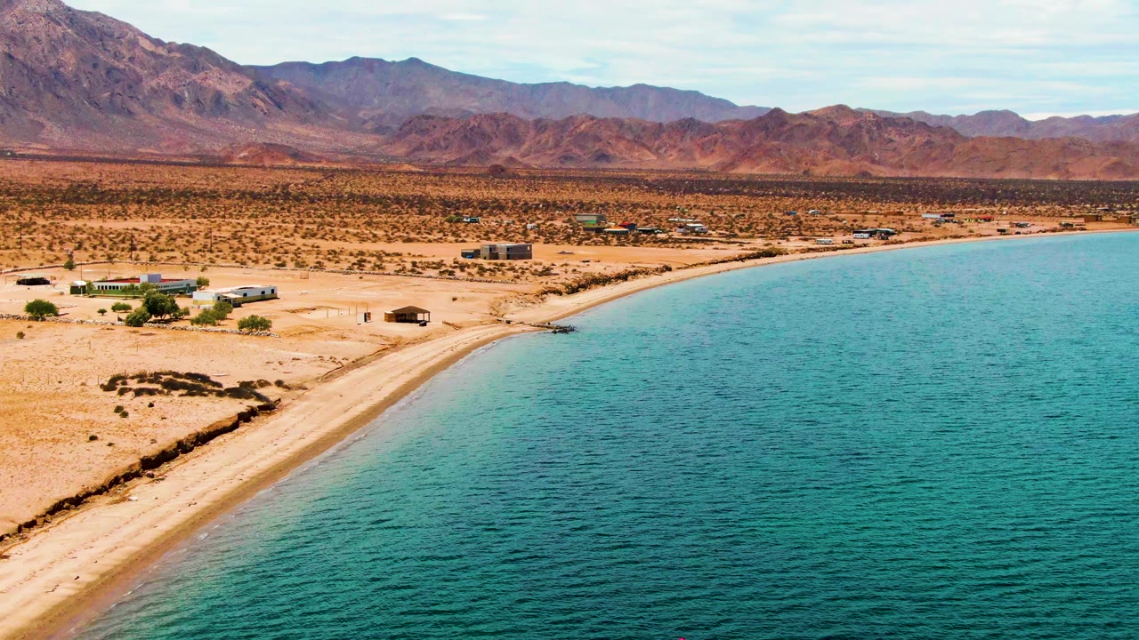 Aerial coastline with sandy desert shore, turquoise water, and mountain horizon.