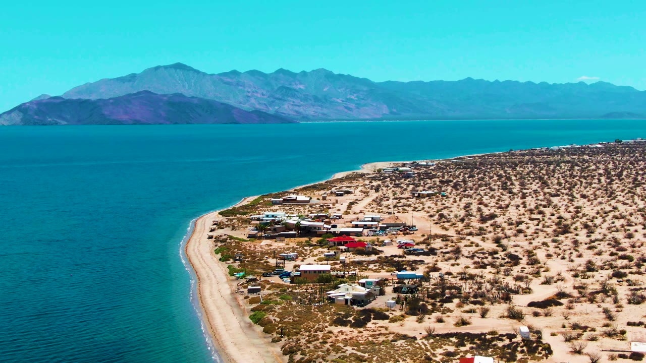 Opening aerial frame over Bahia De Los Angeles with turquoise water on the left, a pale sandy beach curve, desert scrub and small shore structures on the right, distant mountains, and no visible people.