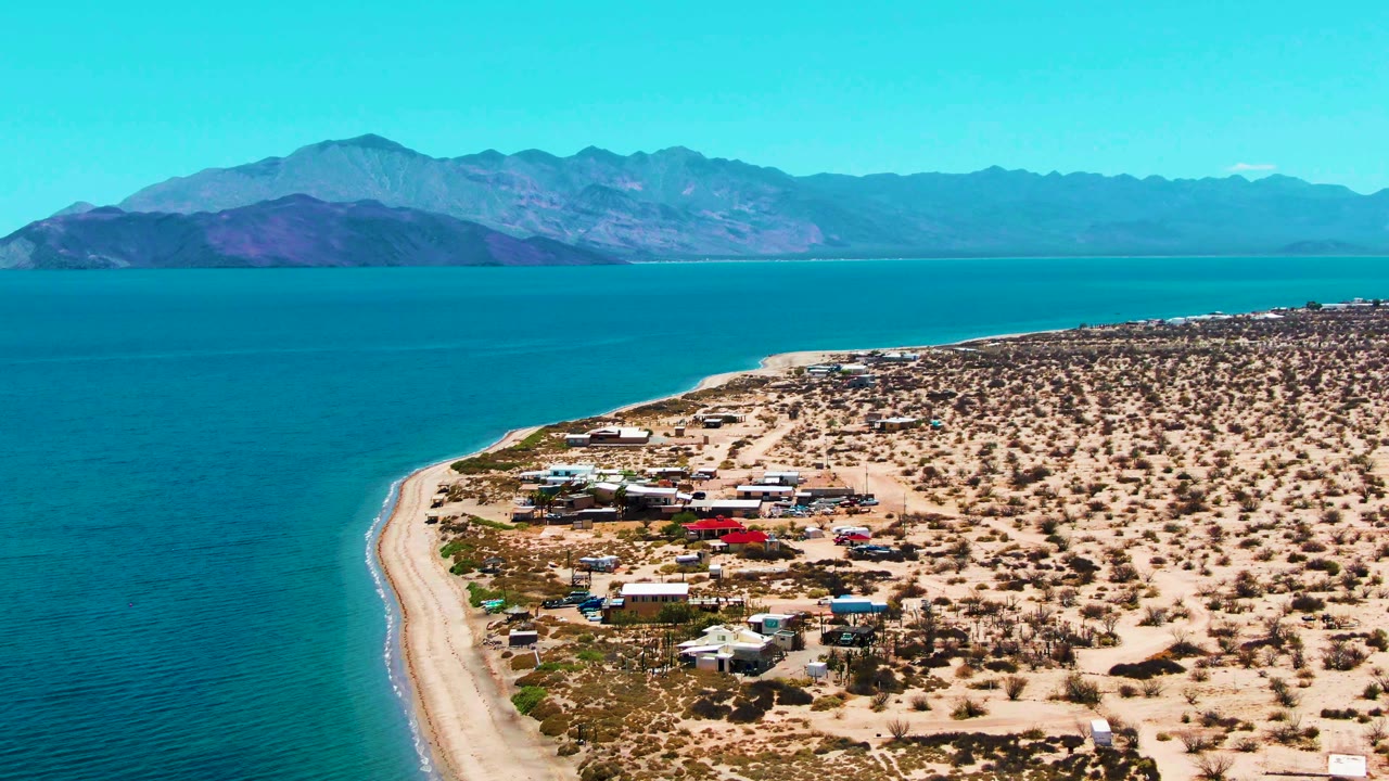 Drone view of a curving beach beside blue ocean and desert buildings under a clear sky.